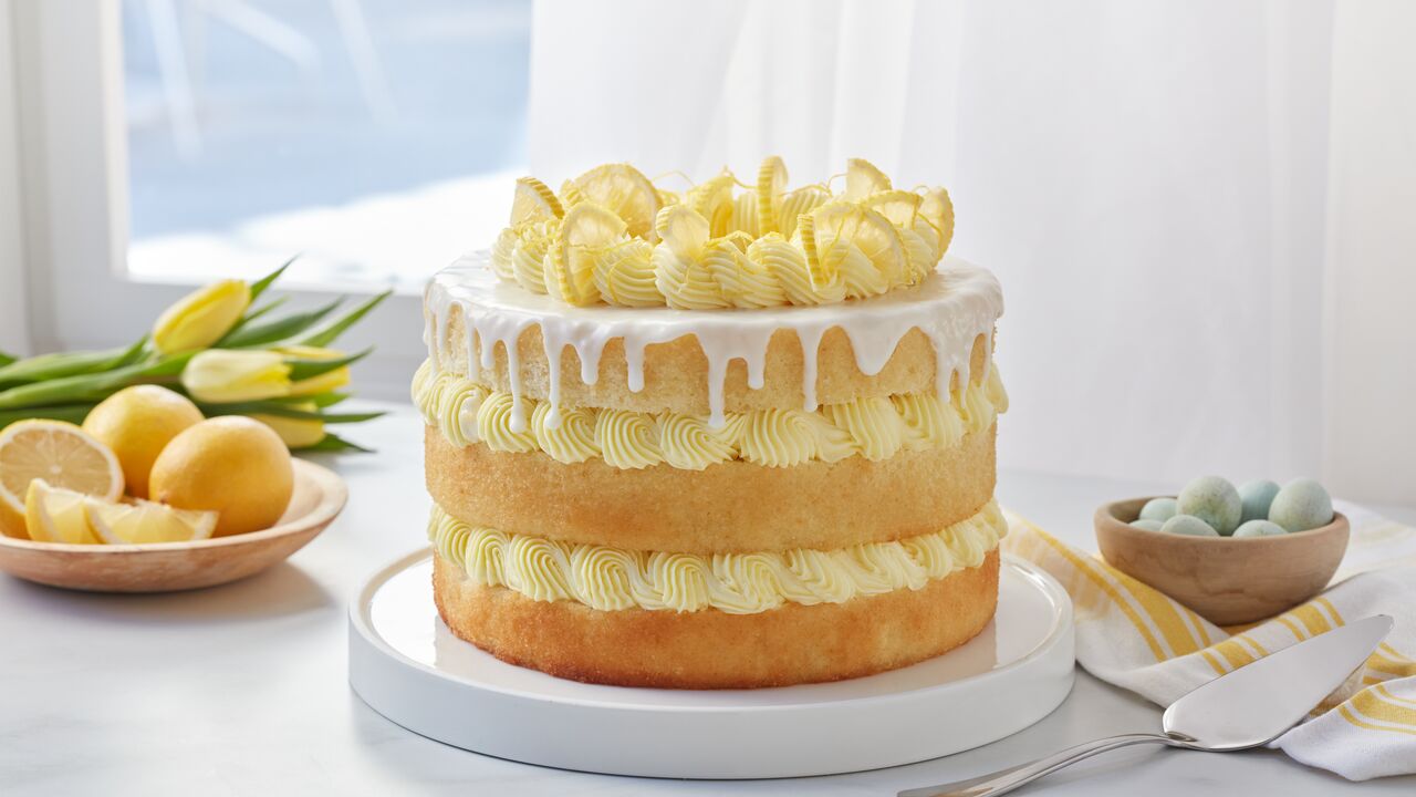 A lemon cake displayed on a counter in front of a window accompanied by a bowl of lemons and a bowl of eggs