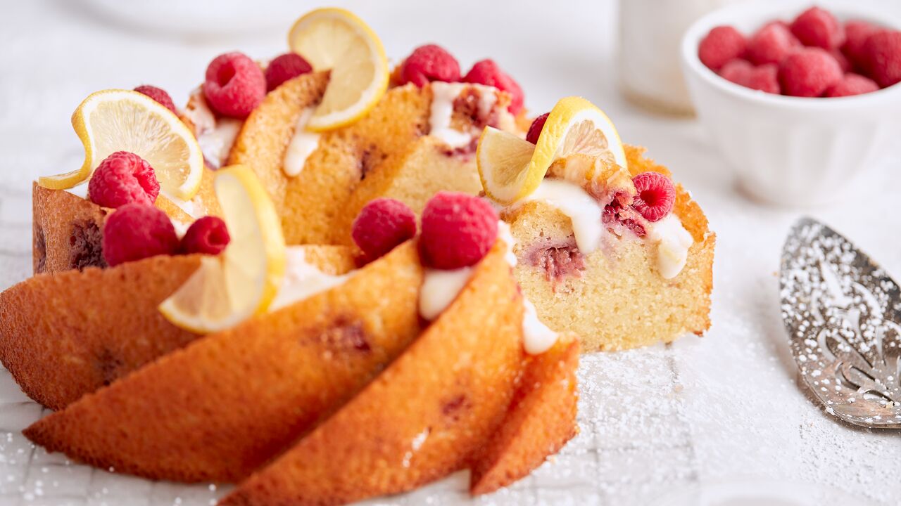 raspberry lemon almond bundt cake displayed on a kitchen counter with fresh raspberries and powdered sugar by Tutti Dolci